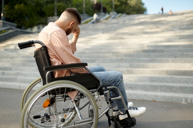 Alone young man in wheelchair at the stairs, handicap problem. Paralyzed people and disability, handicap overcoming. Helpless disabled male person walking in park