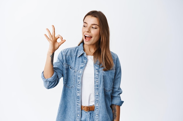 Alright. Smiling cute girl showing okay sign and say yes, approve something good, like and agree, standing pleased on white