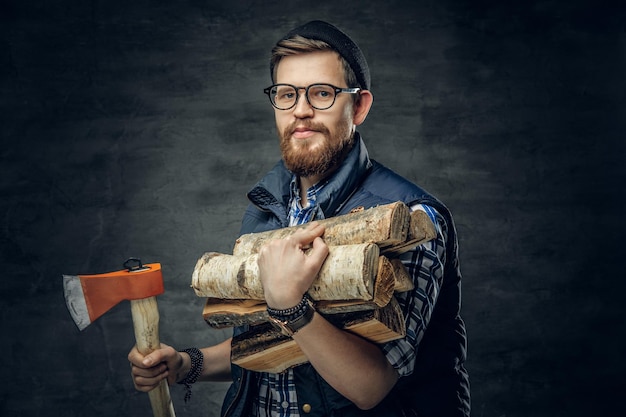 Alternative bearded male in eyeglasses dressed in fleece shirt holds firewoods and axe over grey background.