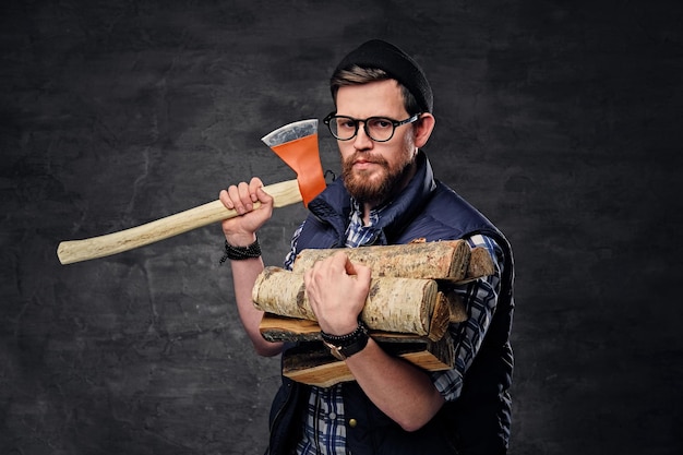 Alternative bearded male in eyeglasses dressed in fleece shirt holds firewoods and axe over grey background.