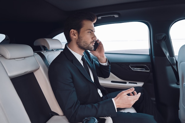 Always available. Handsome young man in full suit talking on the smart phone while sitting in the car