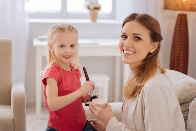 Always beautiful. Cute cheerful positive mother and daughter smiling and looking at you while having a makeup lesson