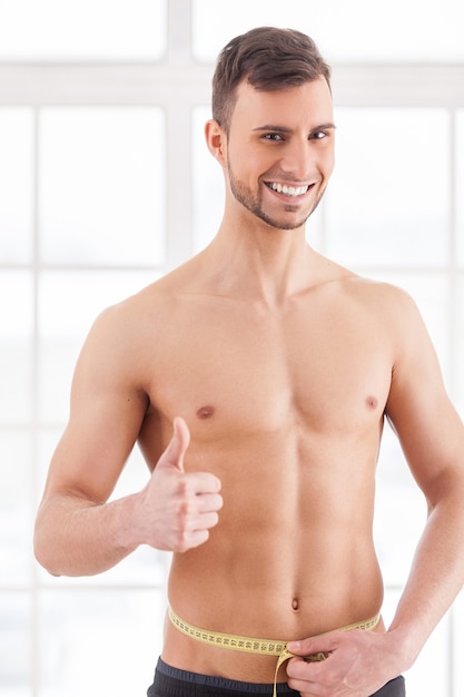 Always in perfect shape. Cheerful young muscular man measuring his waist with measuring tape and gesturing