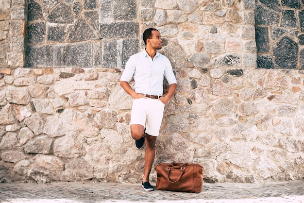 Always in style. Full length of handsome young African man in smart casual clothes standing against the stoned wall outdoors with his bag laying near him