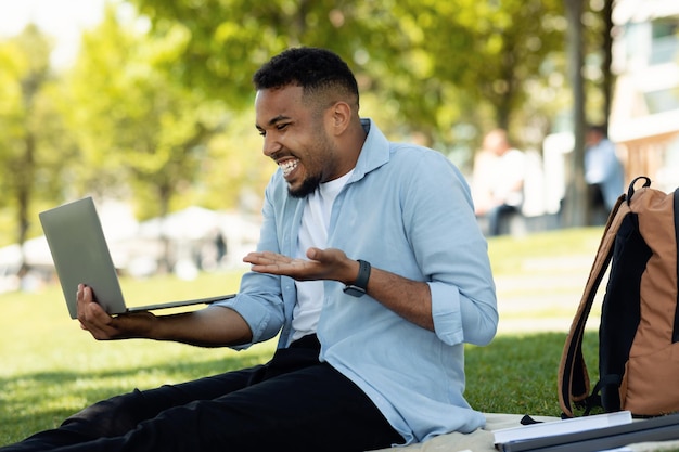 Always in touch concept Excited african american man using laptop outdoors and making video call resting in city park