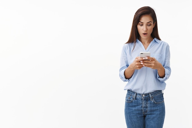 Amazed adult brunette woman read shocking text message chatting friends smartphone app open mouth wondered curiously stare gadget display stand ambushed white background Technology concept