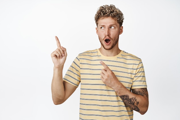 Amazed blond guy smiling while say WOW pointing and looking left with excited and pleased face expression choosing something from shop sale standing over white background