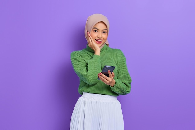 Amazed cheerful beautiful Asian woman in green sweater and hijab using mobile phone isolated over purple background