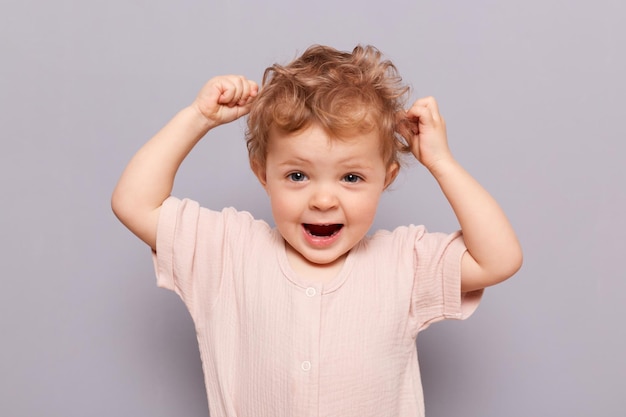 Amazed excited blonde little caucasian infant girl posing isolated over gray background raised arms touching her curly hair screaming with widely opened mouth