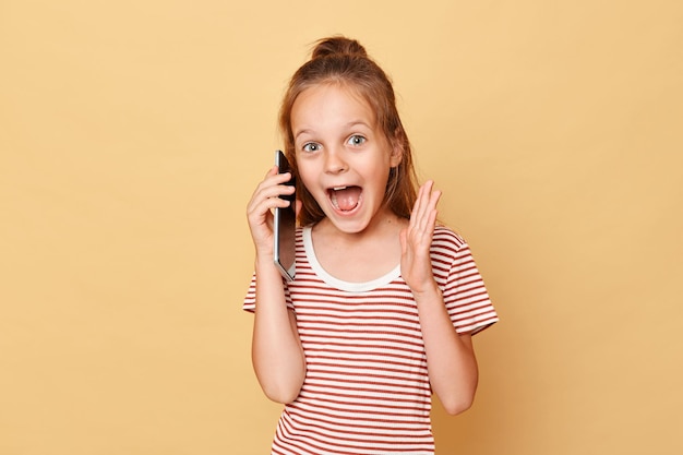 Amazed excited little brown haired girl wearing striped tshirt standing isolated over beige background talking on mobile phone screaming hearing excellent news