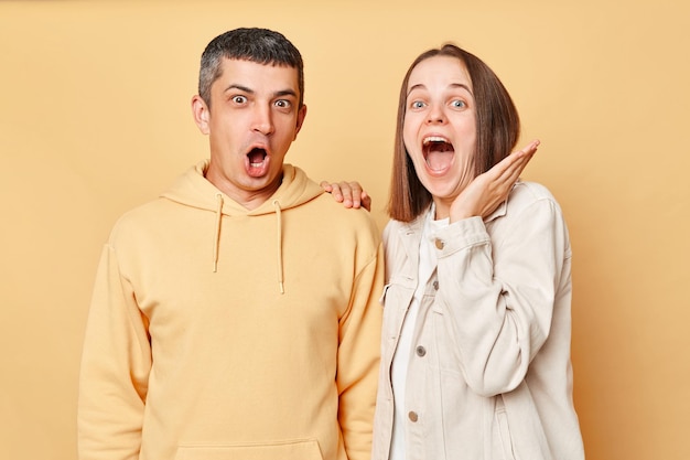 Amazed excited woman and man wearing casual style clothing standing isolated over beige background looking at something astonishing with widely opened mouths