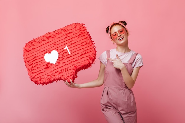 Amazed girl in glasses in shape of hearts holds like sign and looks into camera on pink background