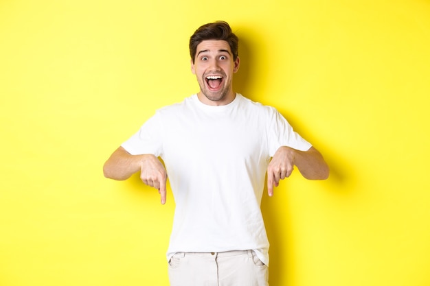 Amazed handsome man pointing fingers down, showing banner with excitement, standing over yellow background