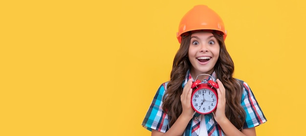 Amazed kid with curly hair in construction helmet hold alarm clock deadline Child builder in helmet horizontal poster design Banner header copy space