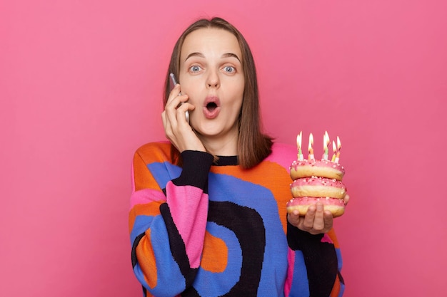 Amazed news on birthday Shocked astonished female in colorful sweater holding delicious sweets with candles talking phone with excited look saying wow isolated over pink background