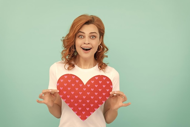 Amazed redhead girl hold red heart on blue background