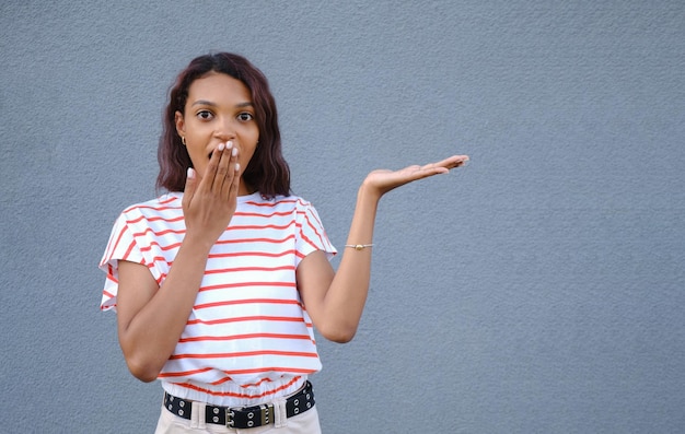 Amazed shocked Afro woman shows good sale offer or deal holds breath demonstrates empty copy space for promo text surprised by big discounts isolated over blue background