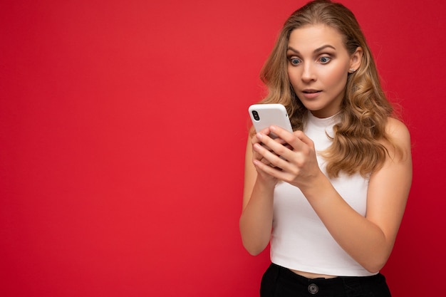 Amazed shocked beautiful young blonde woman wearing white t-shirt isolated over red background using smartphone and texting message via mobile phone looking at gadjet screen