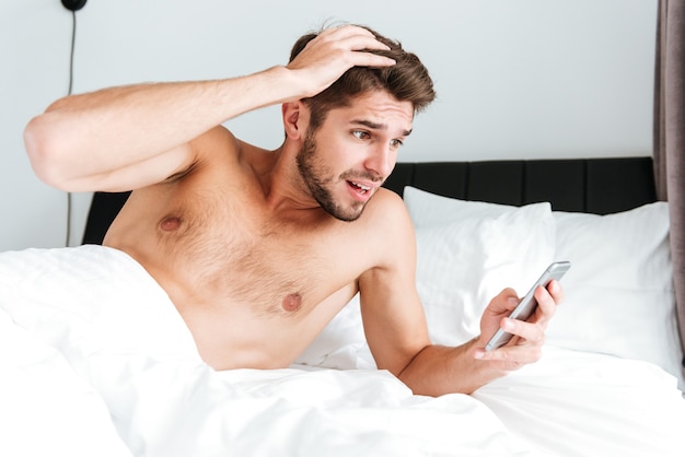 Amazed shocked young man using cell phone in bed in the morning