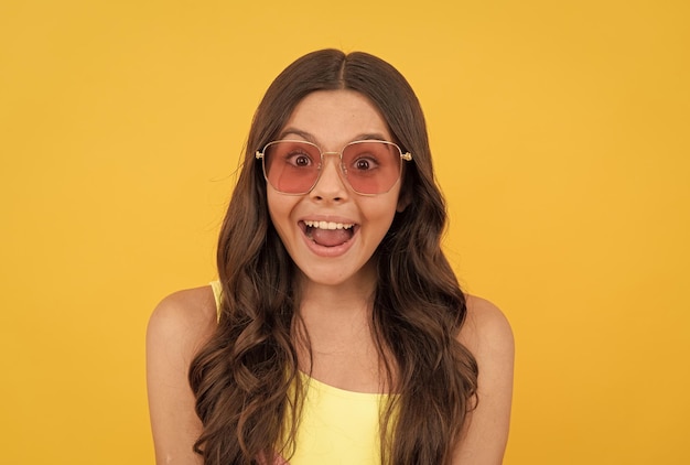 Amazed teen girl in summer glasses has curly hair on yellow background amazement and surprise