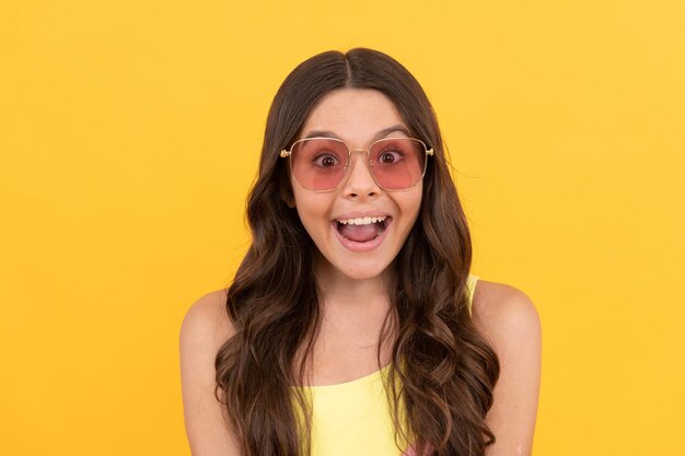 Amazed teen girl in summer glasses has curly hair on yellow background, amazement and surprise.