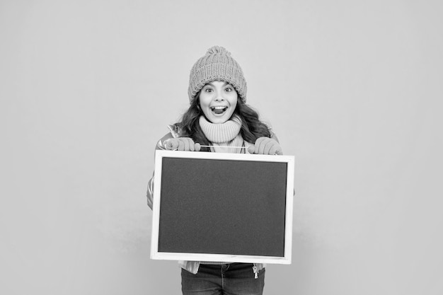 Amazed teen girl in winter clothes hold school blackboard with copy space advertising