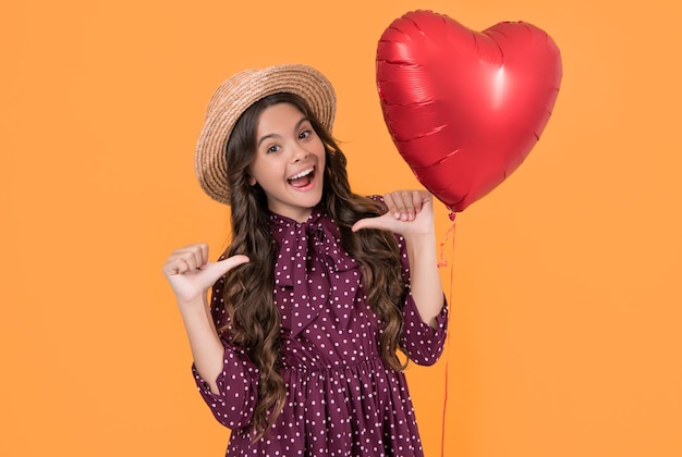 Amazed teen girl with red heart balloon on yellow background point fingers on herself