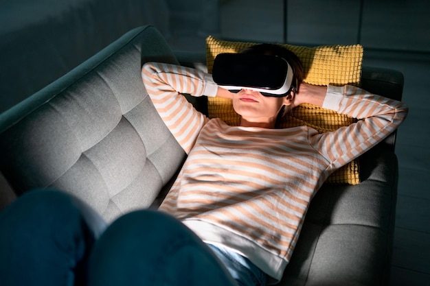 Amazed woman testing modern hightech VR helmet getting 3D experience at home resting lying on sofa