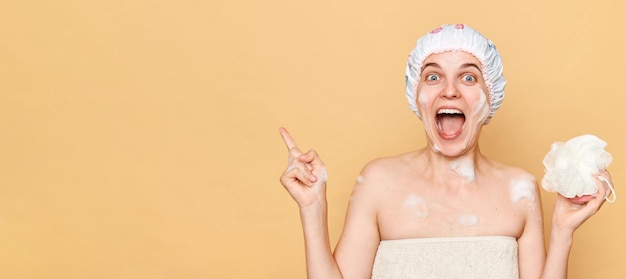 Amazed woman wearing shower cap standing isolated over beige background posing while taking shower pointing at copy space for promotion