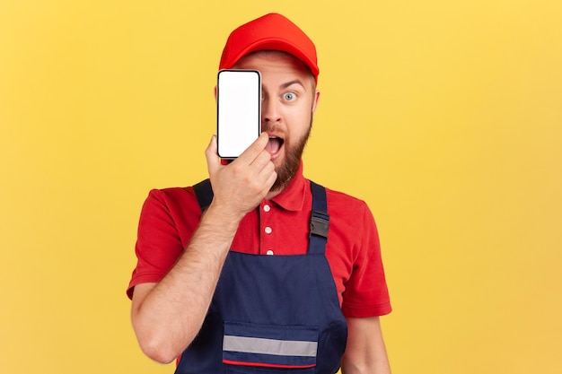 Amazed worker man in blue uniform standing and covering eye with cell phone with empty display