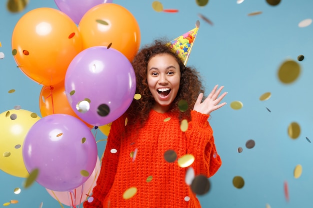 Amazed young african american girl in orange knitted clothes birthday hat isolated on pastel blue wall. Holiday party concept. Celebrating with confetti, colorful air balloons spreading hands.