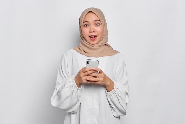 Amazed young Asian Muslim woman using a mobile phone and looking at camera isolated over white background