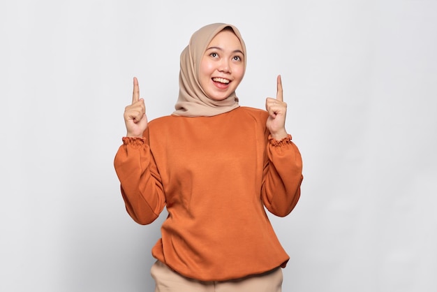 Amazed young Asian woman in orange shirt pointing finger up at copy space isolated over white background