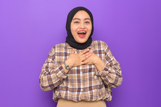 Amazed young Asian woman in plaid shirt hands on chest looking at camera with wow face expression isolated on purple background