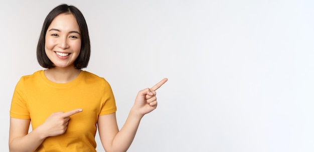 Amazed young asian woman showing advertisement aside pointing fingers right at promotion text brand logo standing happy against white background