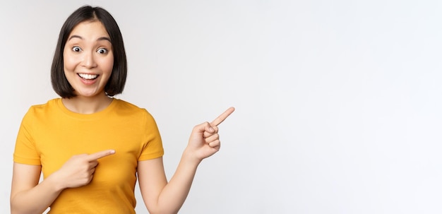 Amazed young asian woman showing advertisement aside pointing fingers right at promotion text brand logo standing happy against white background