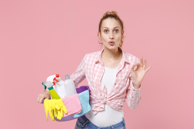 Amazed young woman housewife in checkered shirt hold basin with detergent bottles washing cleansers while doing housework isolated on pink background studio. Housekeeping concept. Showing OK gesture.