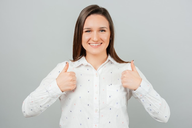 Amazed young woman with toothy smile showing thumbs up gesture over grey background