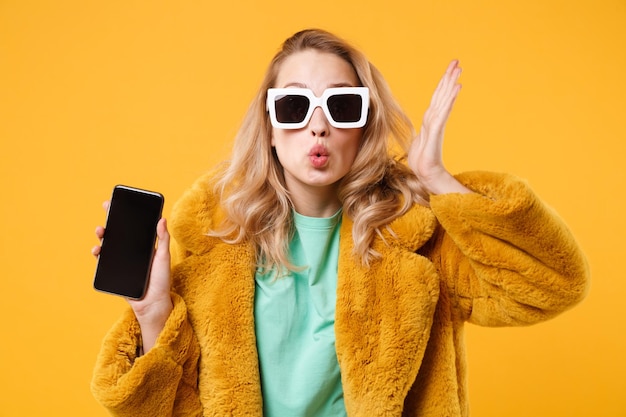 Amazed young woman in yellow fur coat, dark sunglasses posing isolated on orange background. People lifestyle concept. Mock up copy space. Hold mobile phone with blank empty screen spreading hands.