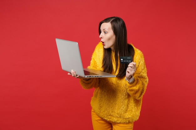 Amazed young woman in yellow fur sweater holding credit bank card, laptop pc computer isolated on bright red wall background in studio. People sincere emotions, lifestyle concept. Mock up copy space.