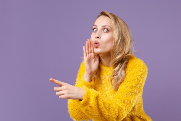 Amazed young woman in yellow fur sweater isolated on pastel violet background. People lifestyle concept. Mock up copy space. Whispers gossip tells secret with hand gesture pointing index finger aside.