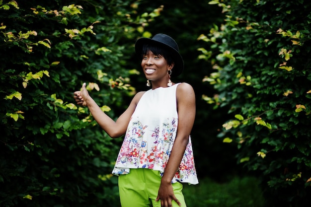 Amazing african american model woman in green pants and black hat posed with different emotions at park shows a thumb up