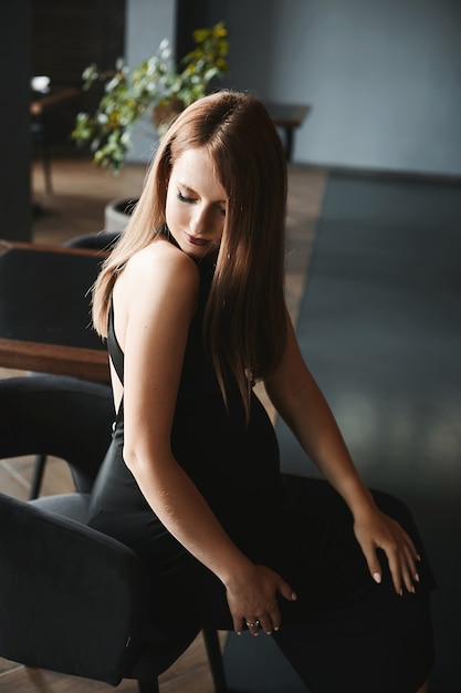 Amazing beauty a young pregnant woman in a trendy black dress is sitting on the chair in the dark in...