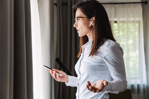 amazing happy beautiful young business woman in formal wear clothes indoors at home talking by mobile phone listening music with earphones using phone.