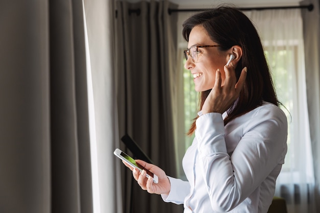 amazing happy beautiful young business woman in formal wear clothes indoors at home talking by mobile phone listening music with earphones using phone.