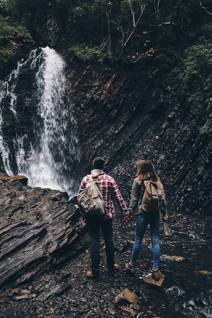 In the amazing place. Full length rear view of young couple holding hands while standing near the waterfall