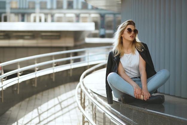 Amazing stylish woman wearing jeans and jacket sitting on street parapet with legs crossed. Copy space