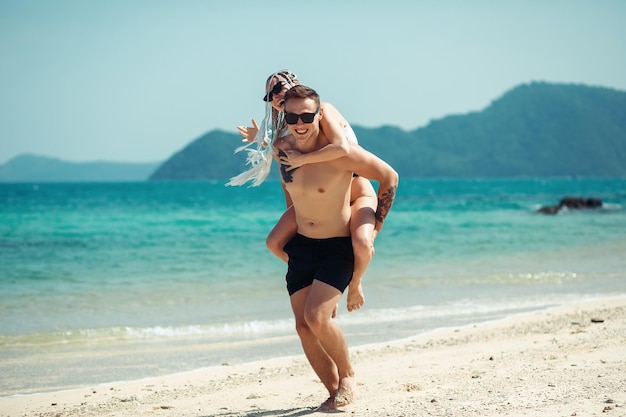 Amazing young couple runs on the beach, the man carries his girlfriend on his back. The young woman with cornrows and in sunglasses laughs; honeymoon concept.