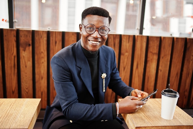 Amazingly looking african american man wear at blue blazer with brooch black turtleneck and glasses posed at street Fashionable black guy with cup of coffe looking at his phone