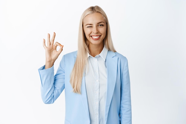 Ambitious corporate woman shows okay sign winks and smiles satisfied at camera approve something good standing in suit over white background